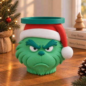 Puede incluir: Un recipiente cilíndrico texturizado de color verde con forma de rostro gruñón, que lleva un gorro de Papá Noel rojo y blanco. El personaje tiene el ceño fruncido, una nariz pequeña y la boca hacia abajo, con un borde verde oscuro.