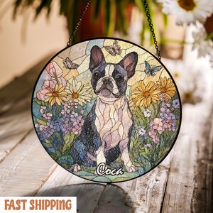 May include: Round stained glass window featuring a Boston Terrier dog surrounded by colorful flowers and butterflies. The dog is black and white, with the name "Coca" written below. The window is hanging by a chain.
