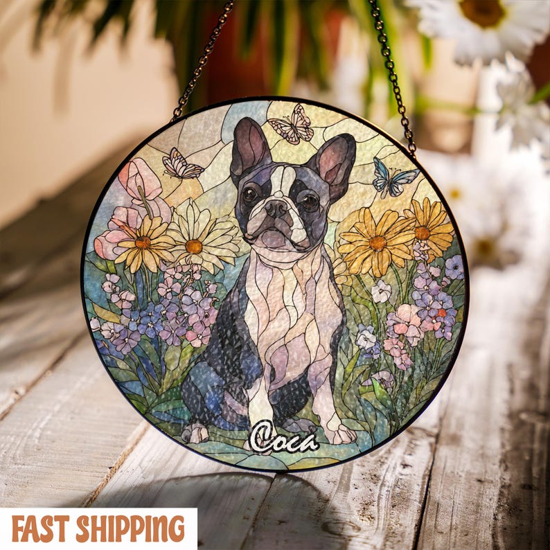 May include: Round stained glass window featuring a Boston Terrier dog surrounded by colorful flowers and butterflies. The dog is black and white, with the name "Coca" written below. The window is hanging by a chain.