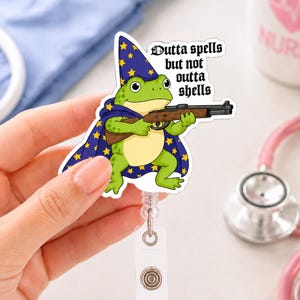 Può includere: Un portabadge con una rana verde che indossa un cappello e un mantello da mago blu con stelle gialle, mentre impugna un fucile. Il testo "Outta spells but not outta shells" è scritto in carattere gotico nero su una base acrilica bianca.