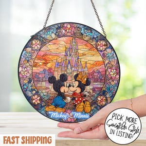 May include: Round stained glass window featuring Mickey and Minnie Mouse kissing in front of a castle. The artwork has a colorful, mosaic-like design with a chain for hanging. The words "Mickey & Minnie" are written on the bottom.