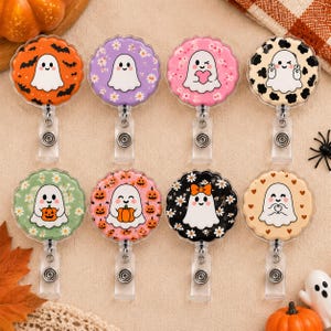 May include: Eight circular badge reels featuring various ghost illustrations. Designs include ghosts with bats, daisies, hearts, cow print, pumpkins, and bows on orange, purple, pink, cream, green, and black backgrounds with scalloped edges.