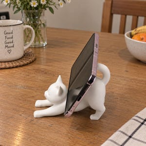 May include: A white cat-shaped phone stand in a stretching pose, supporting a smartphone upright. The cat figure is crafted with a smooth, matte finish and features a curved tail that acts as a support for the device, while the front paws extend forward.