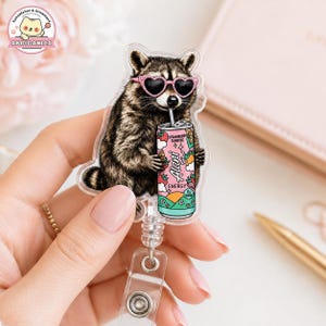 May include: An acrylic badge reel featuring a raccoon wearing pink heart-shaped sunglasses and holding a pink and teal Alani Energy drink can with strawberry and orange illustrations. The design includes the text Strawberry Sunrise and Alani Energy.
