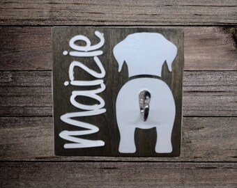 May include: A wooden dog leash hook with the name "Maizie" written on it. The hook is shaped like a dog's rear end and has a silver hook.