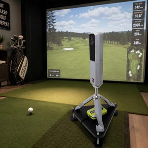 May include: A white Trackman golf launch monitor mounted on a silver tripod base with a black triangular platform. The device stands on a green turf mat near a white golf ball, positioned in front of a large digital screen displaying a golf course.