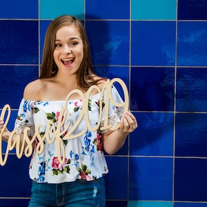 May include: A young woman with long brown hair smiles and holds a wooden sign that says "Class of 2019" in cursive lettering. She is wearing a white off-the-shoulder top with a floral pattern and blue jeans.