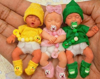 May include: Three miniature baby dolls wearing knitted hats, shirts, and booties. The left doll wears yellow with a duck pacifier clip, the center doll wears pink with a bear pacifier clip, and the right doll wears green with a frog pacifier clip.