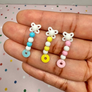 May include: Three handmade beaded earrings with a star-shaped top and a dangling design. Each earring features a different color scheme: blue, yellow, and pink. The earrings are held in a hand.
