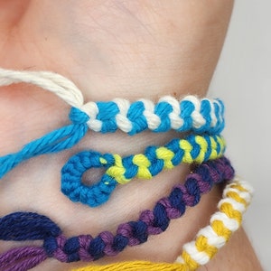 May include: A close-up of a wrist wearing several friendship bracelets in various colors, including white, blue, yellow, purple, and navy blue.