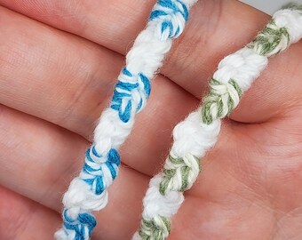 May include: Two handmade friendship bracelets. One is white with blue accents, the other is white with green accents. The bracelets are made of twisted thread and are held in a hand.