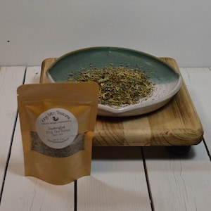 May include: A brown paper bag with a white label that reads "Handcrafted Dog Tea Rinse" and "King Mtn Treasures" with a small chicken illustration. The bag is sitting on a wooden surface next to a teal and white ceramic dish filled with dried herbs.