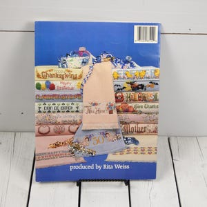 May include: A book cover with a blue background featuring various cross-stitch designs for holidays and special occasions. The designs include text like "Happy Birthday," "Thanksgiving," and "New Home." A decorative towel is draped over the book.
