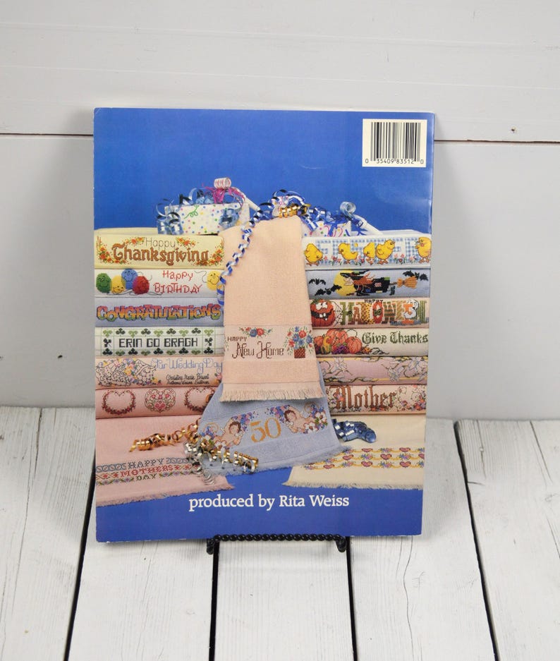 May include: A book cover with a blue background featuring various cross-stitch designs for holidays and special occasions. The designs include text like "Happy Birthday," "Thanksgiving," and "New Home." A decorative towel is draped over the book.