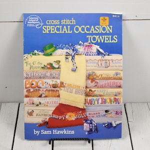 May include: A vintage cross stitch pattern book titled "Special Occasion Towels" by Sam Hawkins. The cover is blue with text and images of cross-stitched towels for various holidays and events, including "Happy 4th of July" and "Happy New Year".