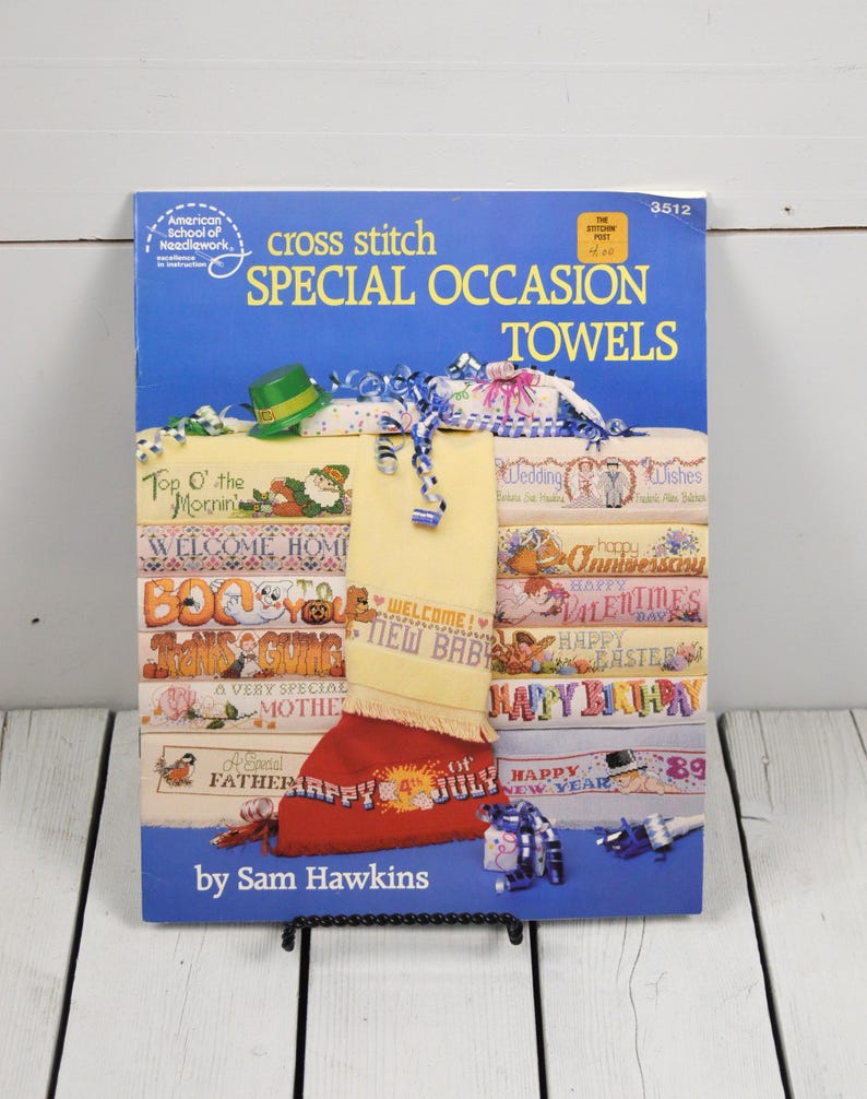 May include: A vintage cross stitch pattern book titled "Special Occasion Towels" by Sam Hawkins. The cover is blue with text and images of cross-stitched towels for various holidays and events, including "Happy 4th of July" and "Happy New Year".