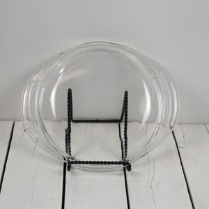 May include: A clear, round glass lid with a smooth surface and integrated side handles. The lid is positioned upright on a black, twisted metal wire display stand, showcasing its transparent design and curved edges for a secure fit.