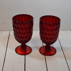 May include: A pair of matching ruby red glass goblets featuring a textured honeycomb pattern on the exterior. Each goblet has a rounded bowl, a short stem with a faceted base, and a circular foot. The glass is translucent and deep red in color.