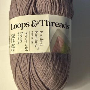 Peut inclure: Pelote de laine Loops & Threads "Brushed Rainbow" de couleur mauve clair. L'étiquette indique qu'il s'agit de polyester, avec 333 m de fil et 150 g. L'étiquette comprend également les traductions anglaises "Brushed Rainbow" et "Polyester".