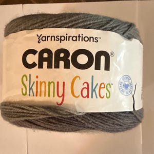 May include: A gray yarn ball with a white label featuring the text "Caron Skinny Cakes" in various colors. The brand name "Yarnspirations" is also visible. The yarn is tightly wound and appears soft.