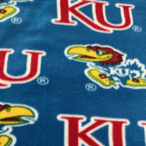 May include: A blue fleece fabric featuring a repeating pattern of the red KU logo and a yellow and red Jayhawk mascot graphic. Each logo and mascot includes a small registered trademark symbol. The texture of the fabric appears soft and fuzzy.