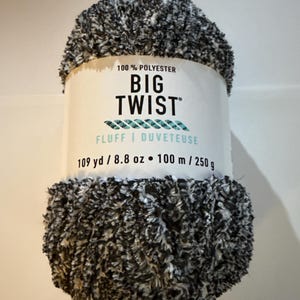 Puede incluir: Ovillo de lana Big Twist, 100% poliéster. El hilo es una mezcla de negro, blanco y gris, con una textura esponjosa. La etiqueta dice "FLUFF | DUVETEUSE" e indica 100 metros / 250 gramos.