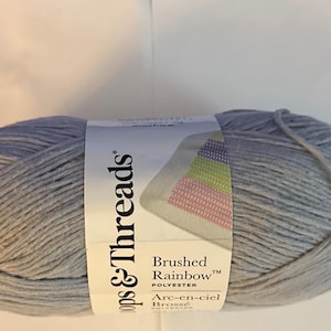 May include: A skein of gray yarn labeled "Loops & Threads Brushed Rainbow Polyester." The label features a colorful geometric design. The yarn is tightly wound and appears soft, ideal for knitting or crocheting projects.