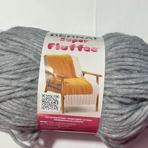 Könnte beinhalten: Ein großer Strang grauer Bernat Super Fluffee Wolle. Das Etikett zeigt einen Stuhl mit einer Decke und den Text "FREE PATTERN ON LABEL". Dieses Garn eignet sich für Strick- und Häkelprojekte.