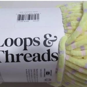 Może przedstawiać: Duża motka przędzy Loops & Threads w miękkim, jasnożółtym kolorze z małymi, równomiernie rozmieszczonymi lawendowymi i różowymi kropkami. Przędza jest owinięta białą etykietą z nazwą marki dużymi, czarnymi literami.