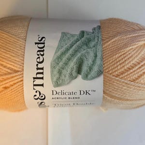 Puede incluir: Un ovillo de hilo Loops & Threads Delicate DK, mezcla acrílica, en color melocotón claro. La etiqueta presenta una manta de punto verde y el texto "Delicate DK Acrylic Blend".