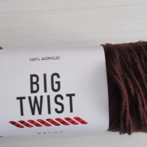 Puede incluir: Un ovillo de hilo Big Twist Value marrón, 100% acrílico. La etiqueta muestra "BIG TWIST" en negrita negra, con un diseño de cuerda roja. El hilo es de un color marrón oscuro.