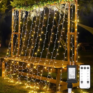 May include: Outdoor string lights with warm white bulbs draped over a wooden bench and arbor. The lights are controlled by a black power adapter and a remote control. The scene is set in a garden.
