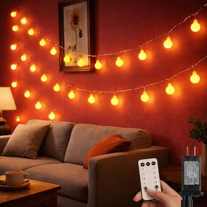 May include: Warm white string lights with round bulbs are strung across a red wall. The lights are above a gray sofa with orange and gray pillows. A remote control is held in a hand. A framed sunflower painting hangs on the wall.