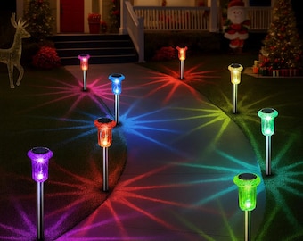 May include: A collection of solar-powered garden lights with colorful light projections. The lights have a silver pole and a colored top, casting red, blue, green, and purple starburst patterns on the pathway and lawn. The scene is set at night with a festive holiday theme.