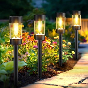May include: A set of four black cylindrical solar path lights with clear glass shades, each featuring a warm glowing filament bulb inside, mounted on thin black stakes positioned along a garden walkway with colorful flowers.