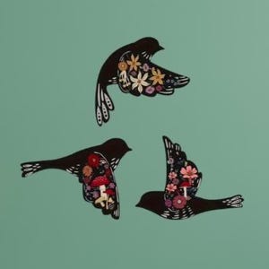 May include: Three black bird-shaped wall decorations featuring intricate cut-out designs on their wings. Each bird displays a colourful internal pattern of various flowers and red mushrooms, creating a detailed contrast against the solid black silhouettes.