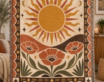 May include: A woven throw blanket featuring a large central sun with wavy rays, set above three stylized orange flowers with green leaves. The design includes a dark brown border with diamond shapes and fringe along the top and bottom edges.