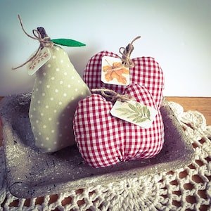May include: A display of fabric fruit, featuring a pear and two pumpkins, presented on a grey tray. The pear is pale green with white spots, and the pumpkins are red and white checked. Each piece is adorned with a small tag.