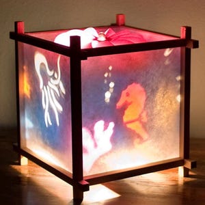 May include: A square lamp with a dark wooden frame and translucent panels featuring illuminated silhouettes of a seahorse, coral, and a jellyfish. The interior light casts a warm glow, highlighting the marine-themed cutouts on the sides.