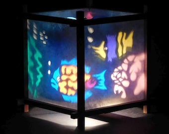 May include: A square lamp with a dark wooden frame and translucent panels featuring a colorful underwater scene. The illuminated panels display vibrant blue, yellow, and green shapes resembling fish, coral, and aquatic plants against a dark background.