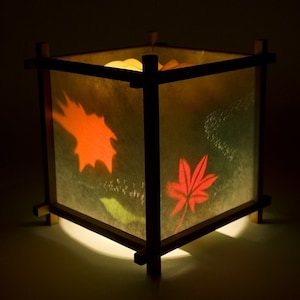 May include: A wooden lampshade with a square shape and a light inside. The lampshade has a translucent paper covering with a design of orange and red leaves. The light shines through the paper, creating a warm glow.