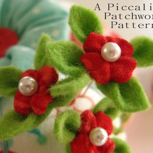 May include: Close-up of a red felt flower with a pearl center, surrounded by green felt leaves. The flower is part of a larger arrangement of felt flowers and leaves, with the text "A Piccalilli Patchwork Pattern" visible in the background.