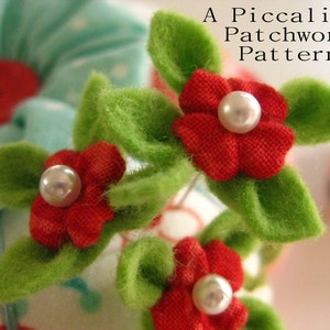 May include: Close-up of a red felt flower with a pearl center, surrounded by green felt leaves. The flower is part of a larger arrangement of felt flowers and leaves, with the text "A Piccalilli Patchwork Pattern" visible in the background.