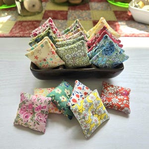 May include: A collection of small, square fabric sachets in a variety of floral and patterned designs. The sachets are arranged in a dark brown rectangular dish and scattered on a white surface. Colors include pink, green, yellow, and blue.