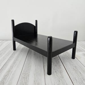 May include: A black wooden doll bed with a simple design. The bed has a headboard and footboard with rounded posts. The bed is made of solid wood and has a smooth finish.