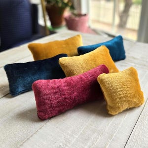 May include: A collection of small, rectangular velvet pillows in various colors, including gold, teal, navy, and magenta. The pillows are arranged on a white wooden surface, showcasing their soft texture and rich hues. These decorative pillows add a touch of luxury.