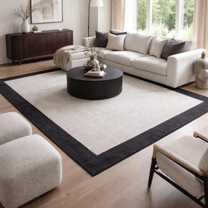 May include: A large rectangular area rug featuring a light beige center panel framed by a wide, solid black border. The rug is positioned on a light wood floor, serving as a central foundation for a living room arrangement with a sofa and coffee table.