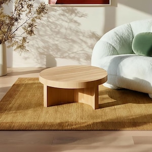 May include: A light wood round coffee table with a simple design sits on a rectangular, golden-brown rug. A white, fluffy armchair with a green pillow is in the background. A white vase with dried branches is on the left.
