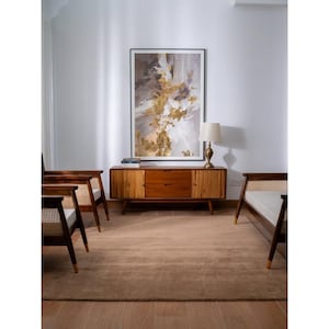 May include: A wooden sideboard with two central drawers and two side cabinets featuring vertical slats, flanked by two wooden armchairs with woven cane backrests and white cushions, topped by a table lamp and a large framed abstract painting.