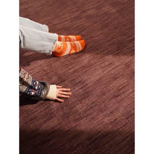 May include: A close-up shot of a textured, deep burgundy rug. The image includes a person's legs in light blue jeans and orange socks with a white pattern. A person's arm in a patterned sweater and a hand is also visible.